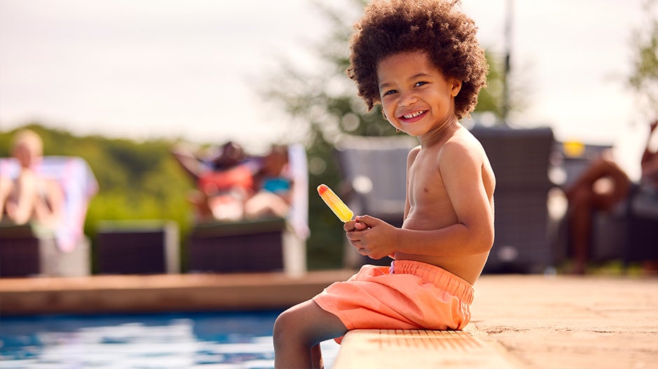 Happy child cooling off with legs in swimming pool, holding popsicle